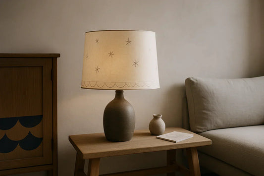 Hand-drawn star lampshade on wood bench with small clay vase, sketchbook, textured beige wall, and gray linen sofa in warm light.