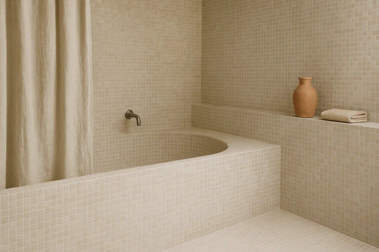 A beige micro-mosaic tiled bathroom with a built-in tub, soft curtain, and a terracotta vase on a ledge.