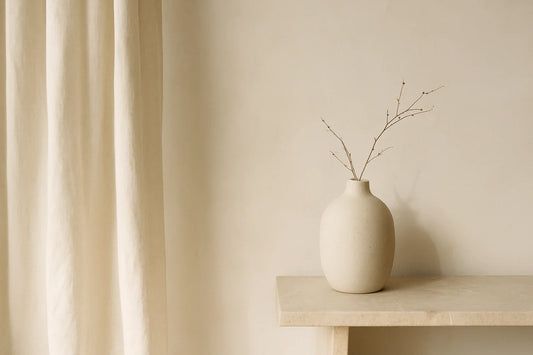 Minimalist room with soft beige walls, linen curtains, and a round ceramic vase holding thin branches on a stone shelf.